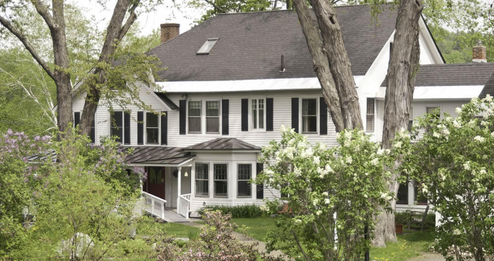 white house with black roof and black shutters surrounded by green lawn and spring blossoming lilacs and trees