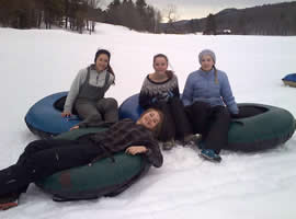 four teen girls sitting on green and blue inner tubes on a tree lined very snowy meadow