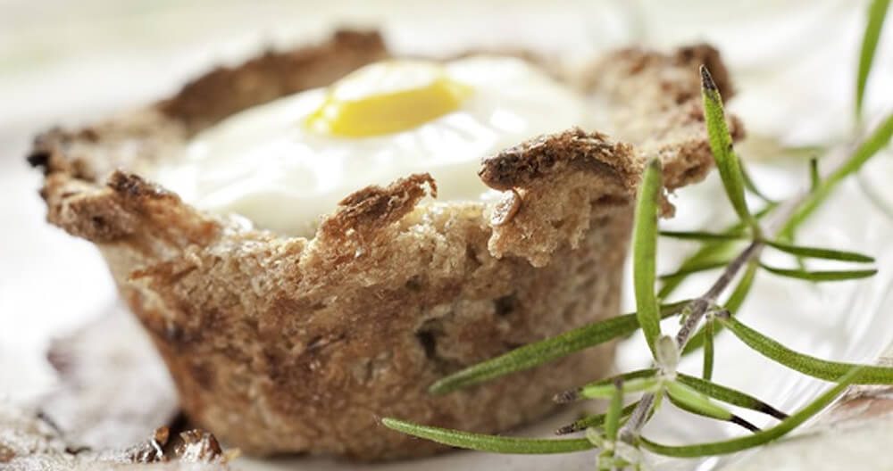 cup made of golden brown bread with white baked egg and yellow yolk center garnished with green rosemary sprig, all on white plate with bacon out of focus in foreground