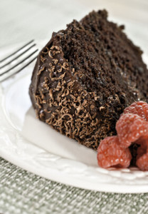 one piece of dark brown chocolate cake with chocolate frosting and shavings laying on its side bright red raspberries all on contrasting white plate with pale green table linen