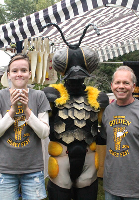 colorful and busy backdrop of vendor tents and signs but in the center of it is two volunteers wearing matching gray and yellow Honey Festival t-shirts standing on either side of a person fully costumed as a honeybee with honeycombed shaped platelets on her belly and a large head with two giant eyes and antennae