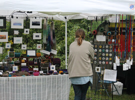 a table full of colorful and varied craft bowls, jewelry, with small framed art hanging on wire screen behind table with one person browsing the merchandise