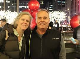 man and woman wearing winter coats posing in front of Christmas decorations with city buildings behind them