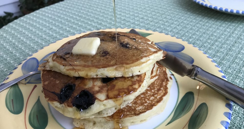 dinner plate with three blueberry pancakes with a pat of butter on top and syrup being poured onto the stack