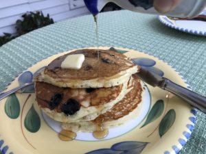 dinner plate with three blueberry pancakes with a pat of butter on top and syrup being poured onto the stack