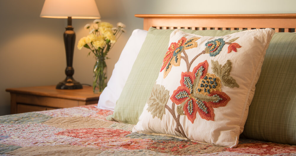 embroidered floral pillow rests against other bed pillows on top of quilted bed with lamp and flowers on end table in background