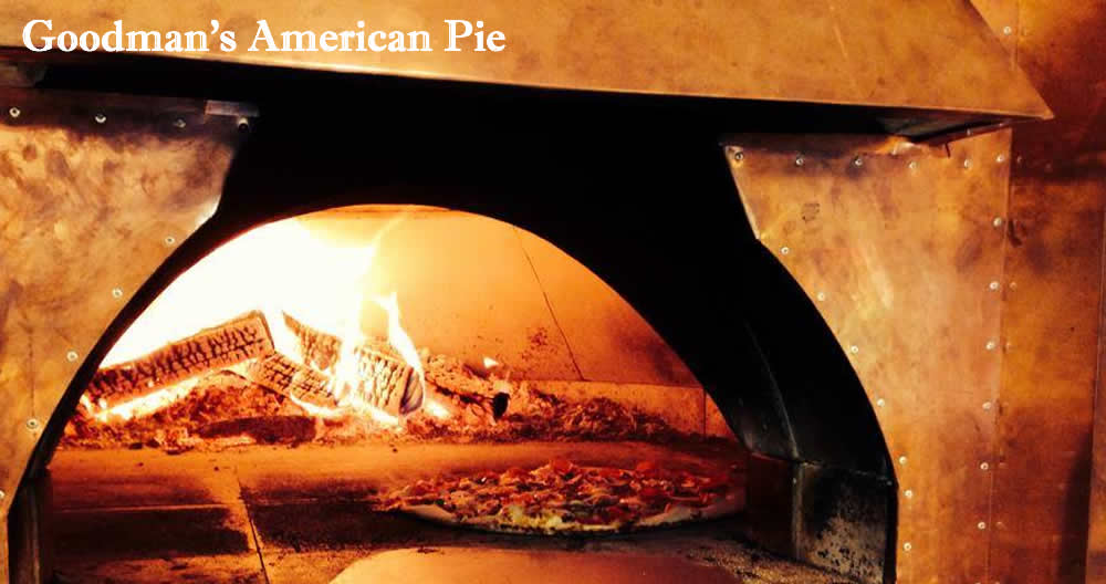 looking into a copper encased wood fired pizza oven, the pizza paddle rests on black flour-covered polished stone in front while deep inside is a pizza baking next to burning logs. The colors are warm and golden.