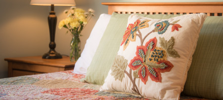 burgundy and gold floral embroideryy on cream colored pillow resting on quilted bed with lamp and vase of flowers in background