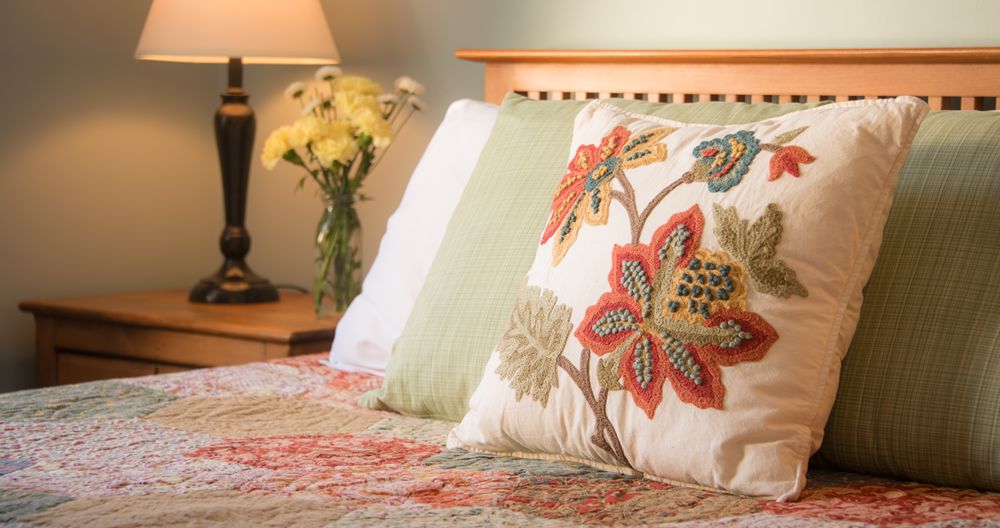 burgundy and gold floral embroideryy on cream colored pillow resting on quilted bed with lamp and vase of flowers in background