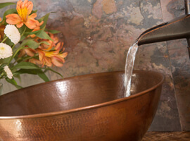 water pouring from antique style faucet into a copper bowl with gray stone tile and vase of orange flowers in background