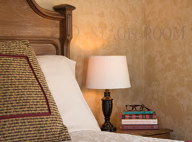 lamp and stack of books with brown reading glasses on a nightstand next to a bed with brown and burgundy pillows and golden brown headboard