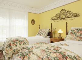 two twin beds side by side with white bedspread and floral comforters at the foot in a bright yellow room with two white-curtained windows