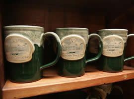 Three green pottery mugs lined up on wooden shelf, each emblazoned with a sketched image of the inn and the name Golden Stage Inn