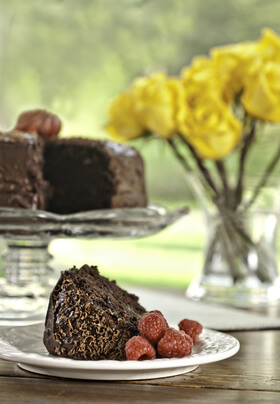 small plate with chocolate cake with red raspberries in front of full cake display and vase of yellow roses