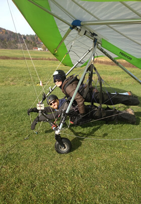 two adults hanging in harnesses under large hang gliding kite frame wearing helmets preparing for flight