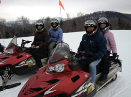 two snowmobiles each with an adult and an older child with helmets surrounded by snowy landscape with hills in the distance