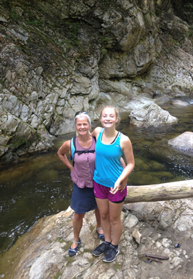 mother and teenage daughter in hiking clothes, looking up at camera, standing on rocky ledge with rocky river set low behind them and a wall of rough rock with woods above them