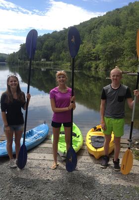 three young adults standing in a row, each with an upright paddle and each with bright blue, green, or yellow kayak behind them, and a tree lined lake with blue sky in the background