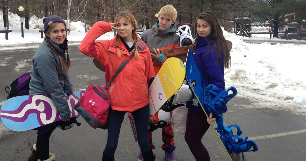 four teenagers with colorful winter coats and boots standing in parking lot with snowboards and gear posing silly for camera