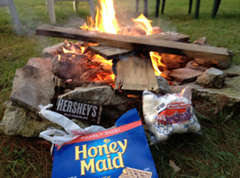 old boards burning in a stone circle campfire with a wrapped hershey’s chocolate bar, a bag full of white marshmallows, and an opened blue box of graham crackers in the foreground